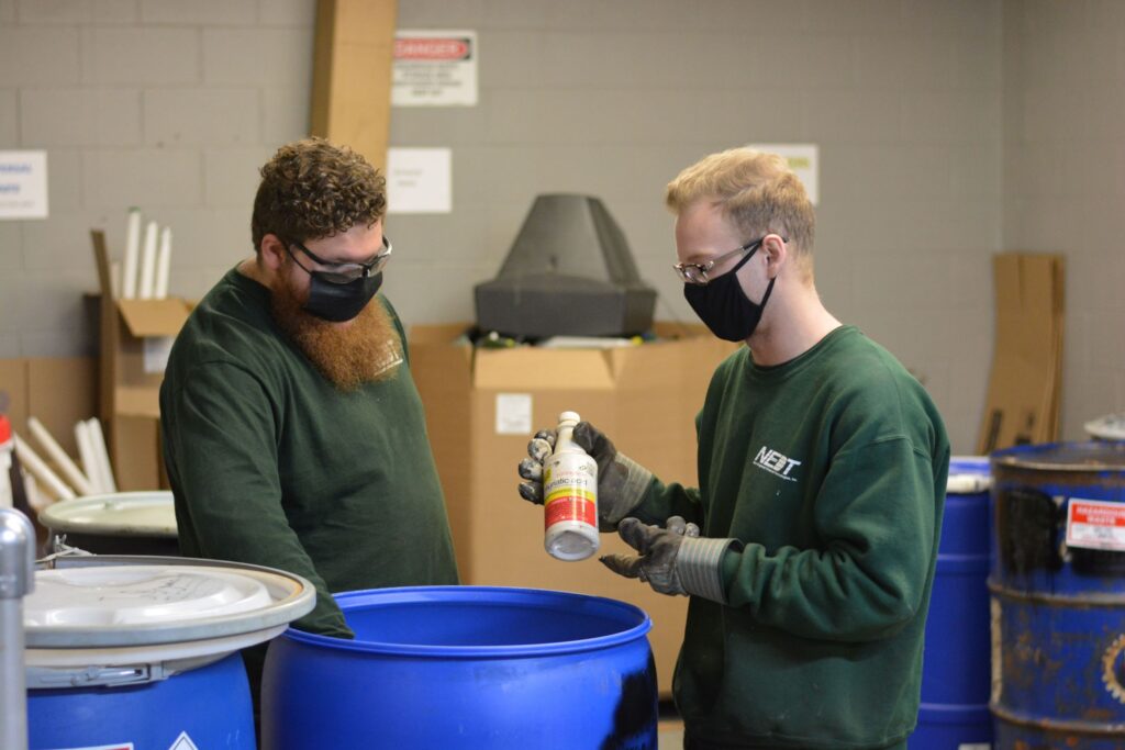 Two NEDT employees hold a bottle of Muriatic Acid above a disposal barrel.