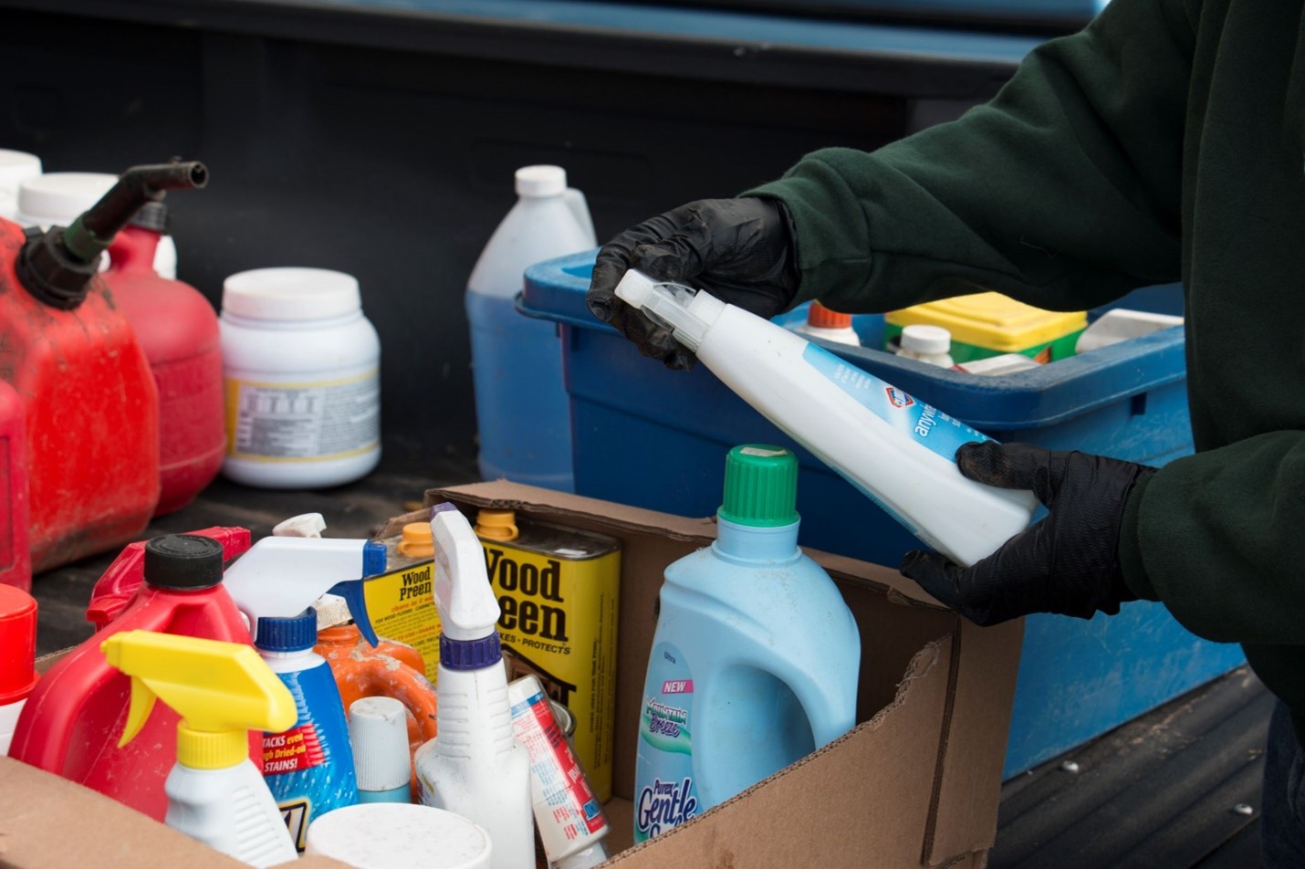 A worker handling a bottle of bleach over a box full of cleaning supplies.