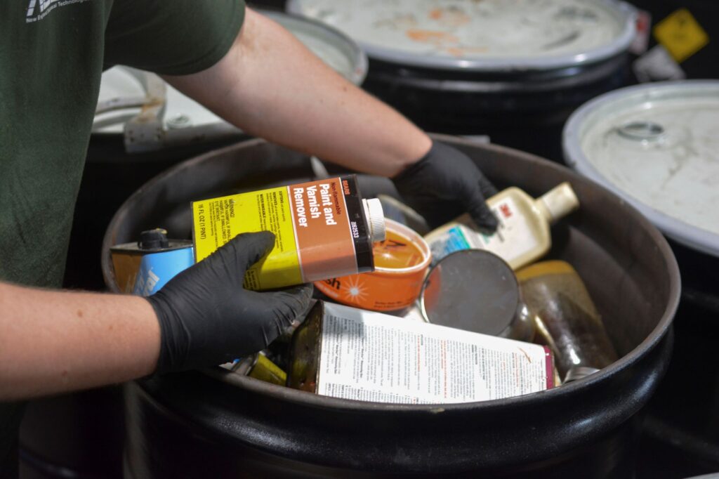 Various household solvents being handled by a NEDT technician for disposal in a barrel.